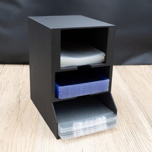 Könnte beinhalten: Ein schwarzes, dreistufiges Aufbewahrungsregal, wahrscheinlich für Karten. Es hat drei offene Fächer, die jeweils einen Stapel klarer oder blauer Kartenhüllen enthalten. Das Design ist einfach und funktional.