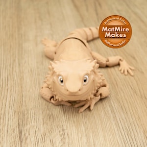 Peut inclure: Figurine articulée de dragon barbu de couleur beige avec une surface texturée détaillée. La figurine a une expression souriante et est posée sur une surface en bois. La figurine porte le texte "MatMire Makes" sur un autocollant circulaire.