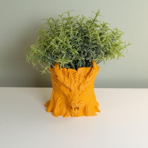 May include: An orange-yellow tree-trunk-shaped planter with a detailed face design, filled with green leafy plants. The planter sits on a white surface, with a light green background. The planter is a decorative item.