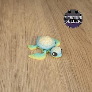 Puede incluir: Una pequeña tortuga de juguete de plástico azul, amarillo y blanco con ojos negros. La tortuga está sobre una superficie de madera marrón claro.