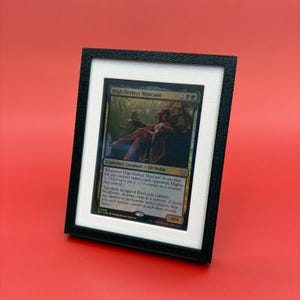 May include: Framed High Perfect Morcant card. The card features a green and blue color scheme with text and an illustration of an elf. The card is framed in a black frame with a white mat.
