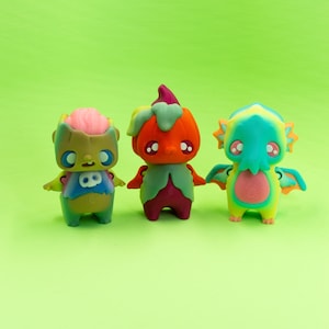 Może przedstawiać: Trzy kolorowe, fantazyjne figurki na zielonym tle. Figurki, w stylu kreskówkowym, przedstawiają zombie, dynię i smoka. Każda z nich ma duże oczy i unikalną kolorystykę, w tym zielony, pomarańczowy i niebieski.