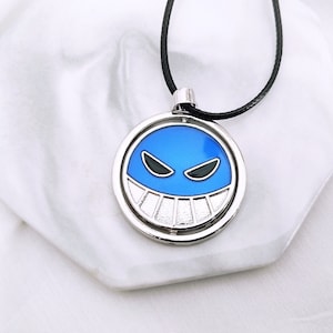 Könnte beinhalten: Runde, silberfarbene Anhängerkette mit blauem und schwarzem Grafikdesign. Der Anhänger zeigt ein lächelndes Gesicht mit scharfen Zähnen und ein schwarzes Band. Die Halskette wird auf einer weißen Oberfläche präsentiert.