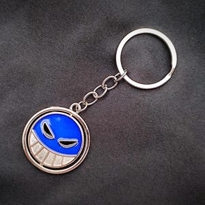 Może przedstawiać: Srebrny brelok do kluczy z okrągłą zawieszką z niebieskim wzorem twarzy z uśmiechniętymi ustami i dwoma kształtami oczu. Brelok ma łańcuszek i okrągły kółko do kluczy.