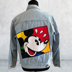 May include: A light blue denim jacket with a colorful Mickey Mouse graphic on the back. The graphic features Mickey Mouse's head with a yellow, red, and blue background.