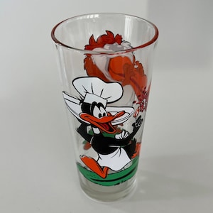 May include: A glass drinking glass with a cartoon image of Daffy Duck dressed as a chef holding a plate and a red rooster with a red and white striped tail. The glass is clear and has a green base.