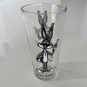 May include: A clear glass drinking glass with a black and white cartoon image of Bugs Bunny. The glass has the words "Bugs Bunny" printed on the bottom.