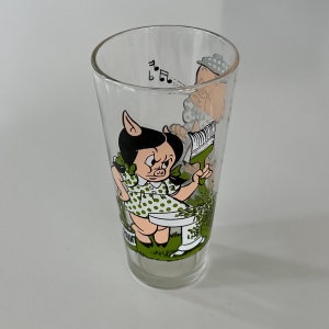 May include: A glass drinking glass with a cartoon image of a pig girl holding a paintbrush and a man with a hat in the background. The glass has green and white accents.