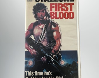 Könnte beinhalten: Ein VHS-Kassettencover mit Sylvester Stallone in einer Militärweste und einem Gewehr. Das Cover ist rot, weiß und blau mit dem Text "Stallone First Blood" und "Diesmal kämpft er um sein Leben!"