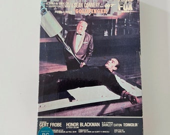 Könnte beinhalten: Ein VHS-Kassettencover für den James-Bond-Film "Goldfinger" mit Sean Connery. Das Cover zeigt eine Szene aus dem Film, in der ein Mann auf einem Tisch liegt, auf den ein Laserstrahl gerichtet ist. Der Text auf dem Cover lautet "CBS FOX VIDEO", "ALBERT R. BROCCOLI and HARRY SALTZMAN present SEAN CONNERY as 007 in IAN FLEMING'S GOLDFINGER", "STARRING GERT FROBE HONOR BLACKMAN", "ALSO STARRING SHIRLEY EATON TECHNICOLOR", "SCREENPLAY BY RICHARD MAIBAUM and PAUL DEHN", "PRODUCED BY HARRY SALTZMAN and ALBERT R. BROCCOLI", "DIRECTED BY GUY HAMILTON", "PARENTAL GUIDANCE PG", und "United Artists".