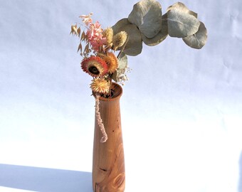 May include: A wooden vase with a flared top contains a display of dried flowers and eucalyptus leaves. The vase is light brown and has a smooth finish. The dried flowers are in shades of orange, pink, and beige.