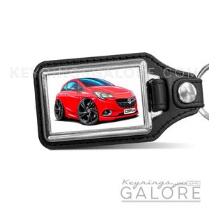 May include: A rectangular black leather keyring featuring a silver metal frame and a central graphic of a red Vauxhall Corsa car with large wheels. The car has a license plate that reads CORSA. The text KEYRINGSGALORE.COM is visible in the background.