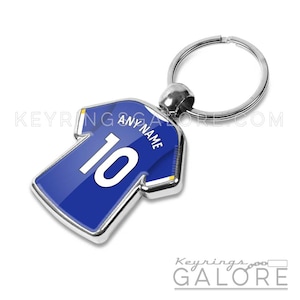 May include: A metal keyring shaped like a blue sports jersey with white trim. The jersey features the number 10 in large white font and the text ANY NAME printed in white above the number. A silver circular split ring is attached to the top.
