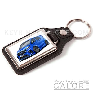 May include: A black leather keychain with a silver-toned rectangular insert featuring a blue Ford Transit van illustration. The van has black wheels and the word "TRANSIT" on the license plate. The keychain has a silver ring.
