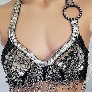 Puede incluir: Top halter negro con adornos plateados. El top presenta un escote con cuentas, detalles de monedas plateadas y cadenas colgantes. Las correas están adornadas con tachuelas plateadas y un gran anillo.