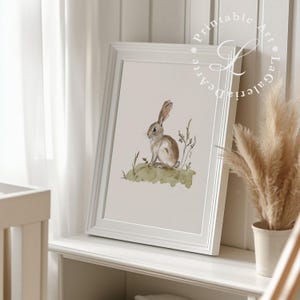 May include: A watercolor painting of a brown rabbit sitting on a green patch of grass. The rabbit is looking to the left of the image. The painting is framed in a white frame.