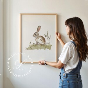 May include: A watercolor painting of a brown rabbit sitting in a field of green grass. The rabbit has long ears and a fluffy tail. The painting is framed in a light wood frame.