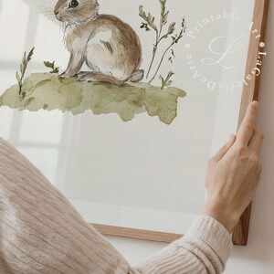 May include: A watercolor painting of a brown rabbit sitting on a green grassy hill. The rabbit is looking to the left and has long ears. The background is a light green watercolor wash.
