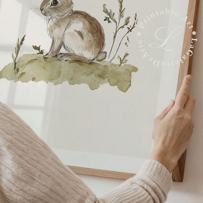 May include: A watercolor painting of a brown rabbit sitting on a green grassy hill. The rabbit is looking to the left and has long ears. The background is a light green watercolor wash.