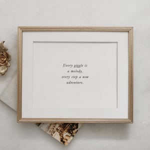 Op de afbeelding: Een gouden lijst met een witte passe-partout en een citaat erin: "Elke giechel is een melodie, elke stap een nieuw avontuur."