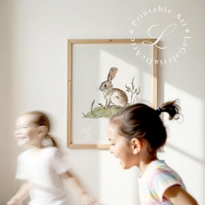 May include: A watercolor illustration of a brown rabbit sitting on a green patch of grass. The illustration is framed in a light wood frame and is hanging on a white wall. Two children are in the background, one is blurred and the other is looking at the artwork.