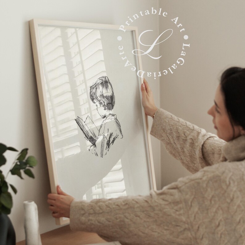 May include: A framed illustration of a child reading a book. The illustration is in black and white and the child is wearing a collared shirt. The frame is white and has a wood-like finish. The frame is being held by a person's hand.