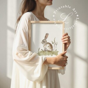 May include: A watercolor painting of a brown rabbit sitting in a field of green grass. The rabbit is looking to the left of the image. The painting is framed in a light wood frame.