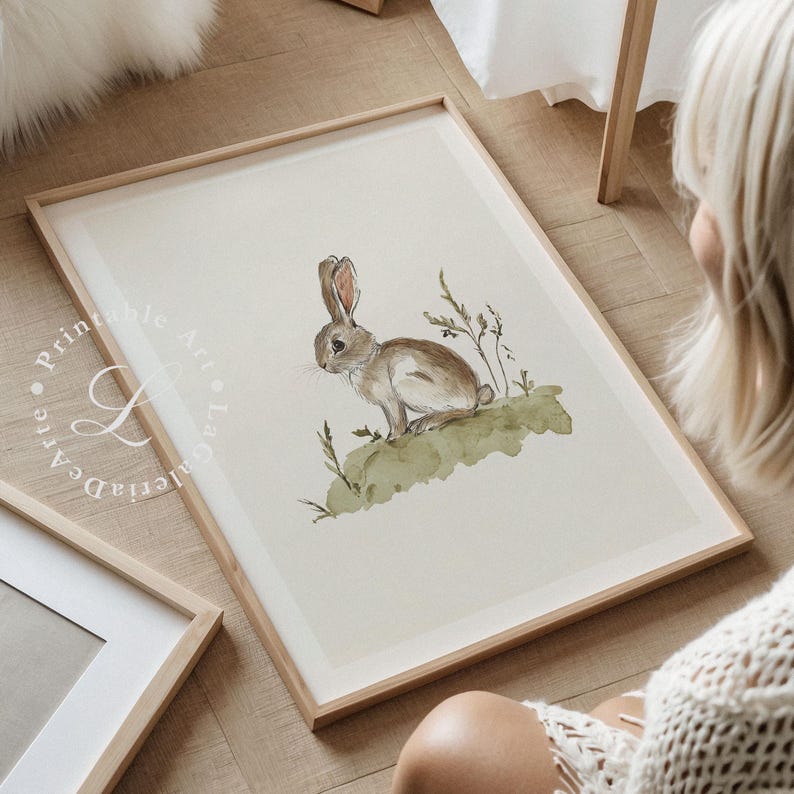 May include: A watercolor illustration of a brown rabbit sitting on a green patch of grass. The rabbit is looking to the left of the image. The illustration is framed in a light wood frame.