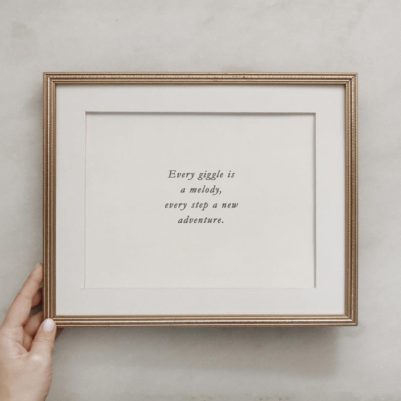 Op de afbeelding: Een gouden ingelijste print met de quote "Elke giechel is een melodie, elke stap een nieuw avontuur."