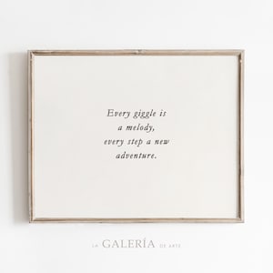 Op de afbeelding: Een ingelijst print met een witte achtergrond en een zwarte tekstquote die luidt "Every giggle is a melody, every step a new adventure." De lijst is van lichtbruin hout.