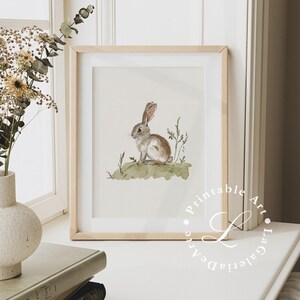 May include: A watercolor painting of a brown rabbit sitting on a green patch of grass. The rabbit is looking to the left of the image. The painting is framed in a light wood frame.