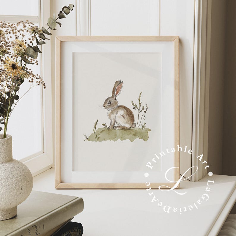 May include: A watercolor painting of a brown rabbit sitting on a green patch of grass. The rabbit is looking to the left of the image. The painting is framed in a light wood frame.