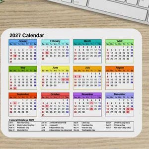 May include: A rectangular 2027 calendar mouse pad featuring twelve monthly grids with color-coded headers. Each month displays dates in black text with specific dates highlighted in red. The bottom section lists Federal Holidays 2027 in black text.