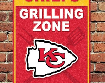 Pode incluir: Um sinal de metal vermelho e amarelo com as palavras "Chiefs Grilling Zone" em branco. O sinal apresenta o logotipo do Kansas City Chiefs e o texto "Seja um jogador de equipe, mantenha nosso mestre do churrasco hidratado."