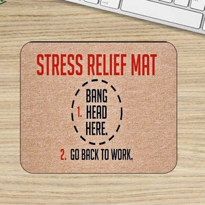 May include: A brown desk mat with the text "Stress Relief Mat" in red. The mat has instructions: "1. Bang Head Here." and "2. Go Back To Work."