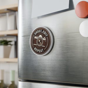 Könnte beinhalten: Ein runder brauner und weißer Magnet mit einer Vintage-Kamera-Illustration und dem Text "Shoot, Edit, Repeat".