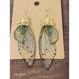 May include: Dangle earrings with a butterfly wing design. The earrings have gold-coloured filigree tops with green and gold accents. The wings are translucent with gold flecks and black vein detailing.