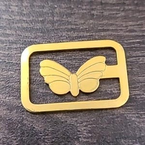 May include: A gold-colored metal bookmark featuring a butterfly design within a rectangular frame. The butterfly has detailed wing patterns. The bookmark is designed to slide over a page.