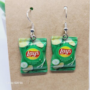 Puede incluir: Un par de pendientes novedosos con miniaturas de bolsas de patatas fritas Lay's Sour Cream & Onion verdes y blancas. Los pendientes tienen ganchos para las orejas de color plateado y el logotipo de Lay's es visible.