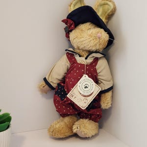 May include: A plush tan rabbit toy wearing a dark blue hat with a red polka dot bow and red overalls featuring a white star pattern. The rabbit has a pink nose and a beige shirt. A paper tag attached to the overalls reads BE CAUTIOUS SAYS THE HEAD BEAN.