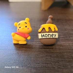 May include: Two small, colorful figurines. One is a yellow bear with a red shirt and pink cheeks. The other is a brown honey pot with yellow honey dripping down and the word "Honey" on a white label.