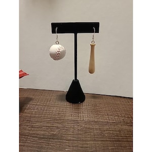 Op de afbeelding: Oorbellen met honkbalthema. Eén oorbel is een witte honkbal met rode stiksels, de andere is een lichtbruine honkbalknuppel. De oorbellen hangen aan zilverkleurige haken.