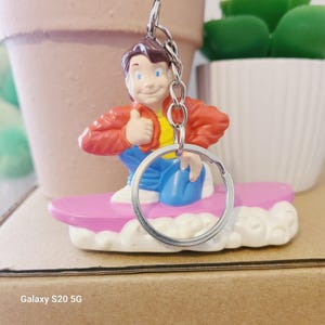 May include: A novelty keychain featuring a cartoon character in a red jacket, yellow shirt, and blue pants, riding a pink surfboard. The character is giving a thumbs-up. The keychain has a silver metal ring.