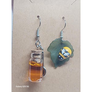 May include: A pair of unique earrings. One earring features a small glass bottle filled with amber liquid and a cork stopper. The other earring has a green leaf with a yellow and black bee design. Both earrings have silver-colored hooks.