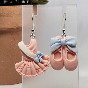 May include: A pair of mismatched dangle earrings featuring a light pink ballet dress with a white sash and small blue bow on one side, and a pair of light pink ballet slippers topped with a large light blue bow on the other, with silver hooks.