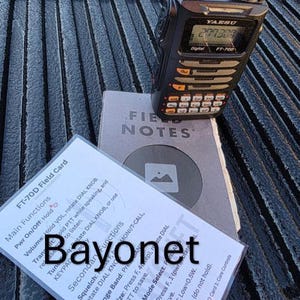 Könnte beinhalten: Ein schwarzes und graues Yaesu FT-700 Digitalfunkgerät mit Tastatur und Display, das auf einem Notizbuch und einer Feldkarte liegt. Auf der Feldkarte steht das Wort "Bayonet".