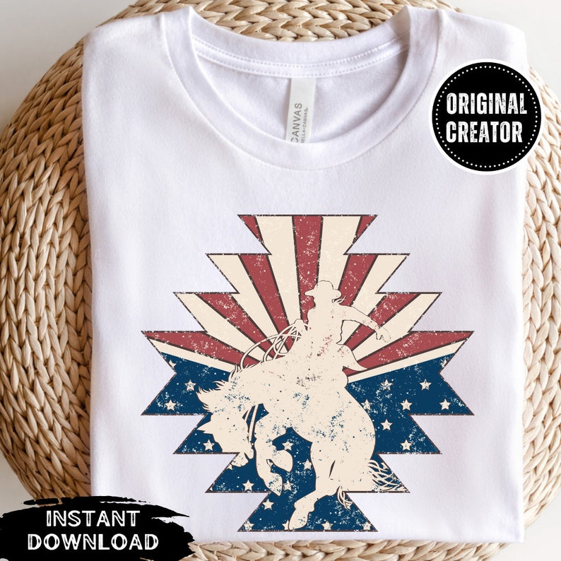 May include: A white t-shirt with a distressed vintage design of a cowboy riding a horse in front of a red, white, and blue American flag. The design is in a southwestern style with a starburst pattern.