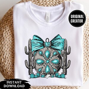 May include: A white t-shirt featuring a turquoise and grey Christmas bauble design. The bauble is surrounded by two cacti and a turquoise bow. The text "Original Creator" is in a white box with a black border.