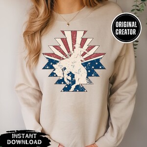 May include: A beige sweatshirt with a vintage-style graphic of a cowgirl riding a horse in front of a red, white, and blue American flag design.