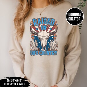 Pu&ograve; includere: Una felpa beige con un design grafico di un teschio di mucca con il testo "Raised on 90's Country" in blu e rosso.